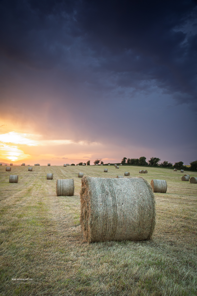 Foreground hay bale beneath dark storm clouds and a glowing sunset break near Gort in County Galway