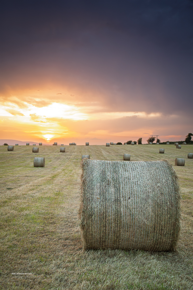 Large round hay bale in a harvested field under golden sunset light and receding storm clouds near Gort, County Galway.
