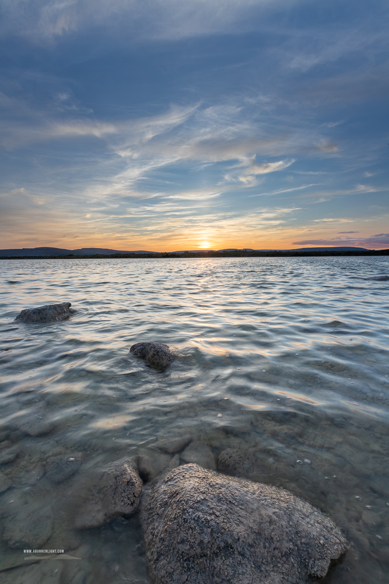 A limestone boulder rises from rippled blue water beneath a sweeping summer sunset sky at Lough Bunny, County Clare.